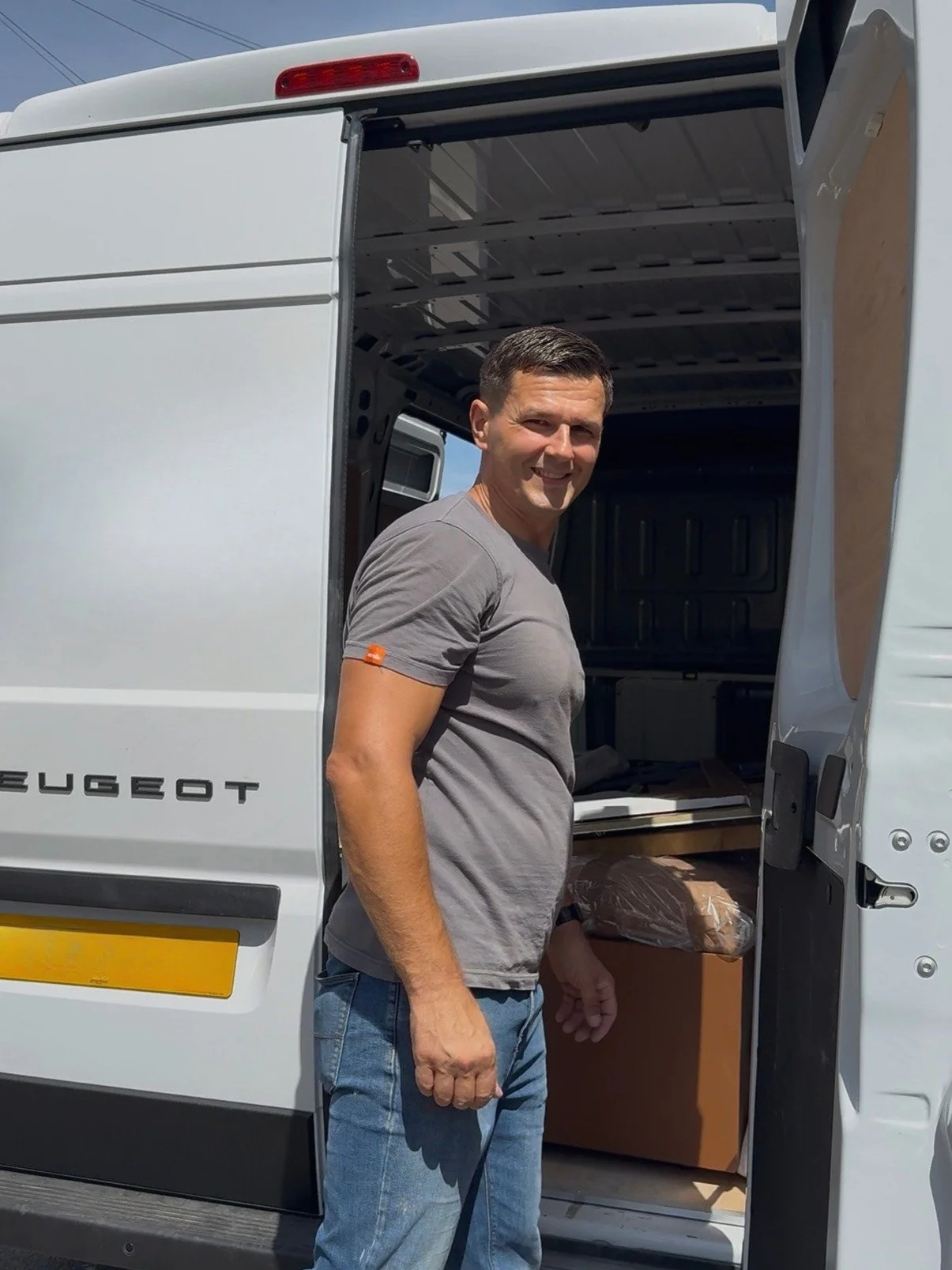 A man standing next to a white delivery van with the rear door open, revealing boxes inside, outdoors on a sunny day.