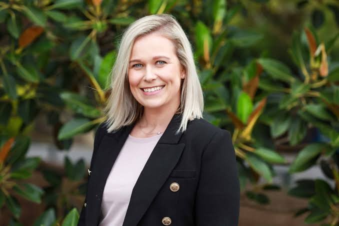 A young woman with blonde hair smiling at the camera, wearing a black blazer and standing outdoors with green foliage in the background.