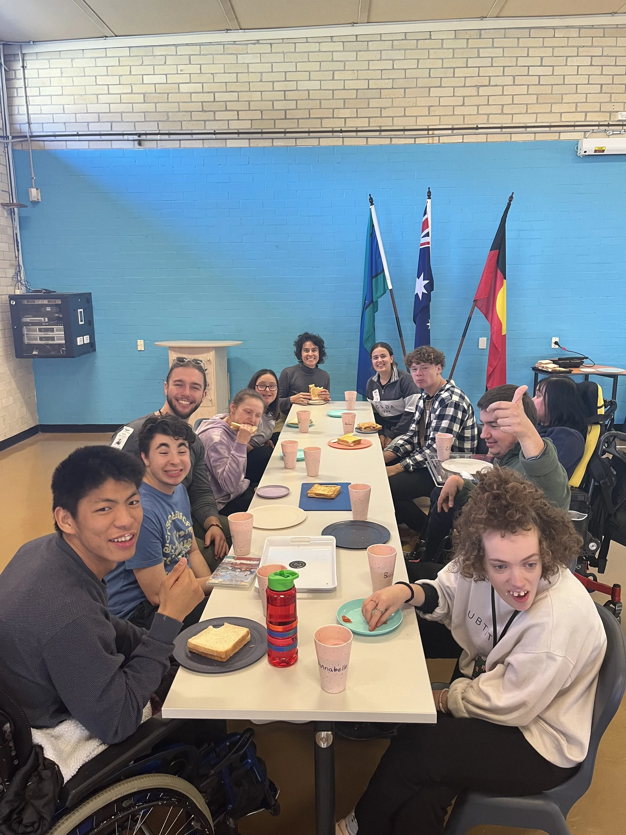 Group of young people, some in wheelchairs, sitting around a long table in a bright room. They are smiling and enjoying snacks like toast and drinks. In the background, there are three flags on stands against a blue wall with a brick pattern. The room has a high ceiling with visible pipes.