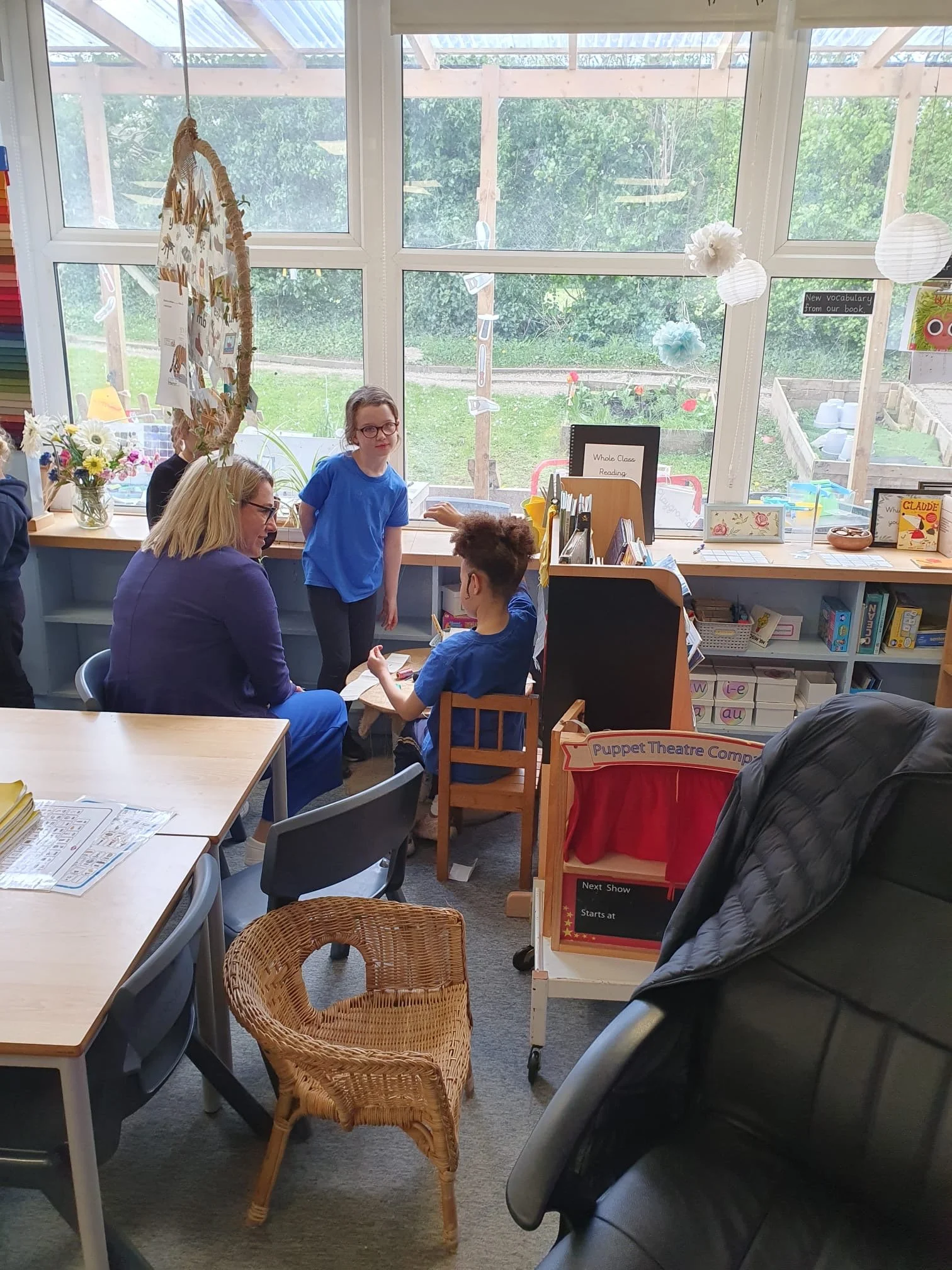 A classroom with large windows showing a green outdoor area. Inside, a woman with glasses and long blonde hair, a boy with glasses, and a girl with curly hair are engaged in activities. There are various educational materials, books, and decorations, including paper lanterns and a sign that reads 'Whole Class Reading.' A basket chair is in the foreground, and the room is well-lit with natural light.