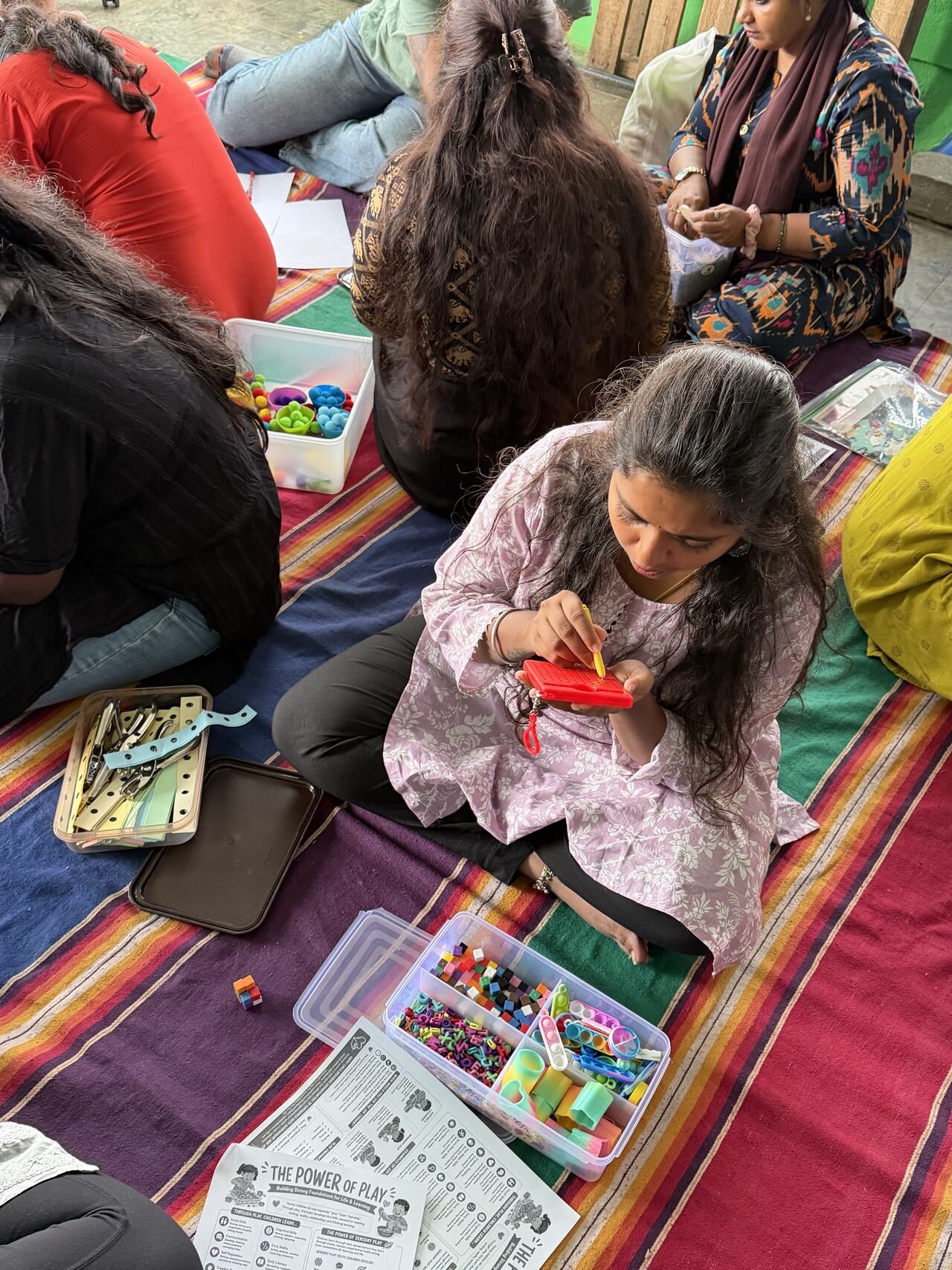 Group of women sitting on a colorful striped mat, engaged in arts and crafts using various craft supplies such as beads, clips, and string, with papers and containers around them.