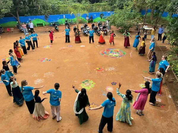 A group of children and adults participating in a traditional Indian dance circle outdoors, with colorful rangoli decorations on the ground, trees, and a blue fence in the background.