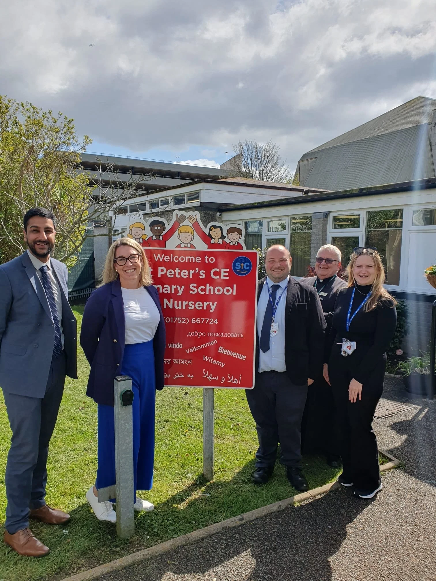 A group of five people standing outdoors next to a red sign at a school event. The sign reads "Welcome to St. Peter’s CE Primary School Nursery" in multiple languages. The group is dressed in business and casual attire, smiling for the camera with trees and a building in the background under a cloudy sky.