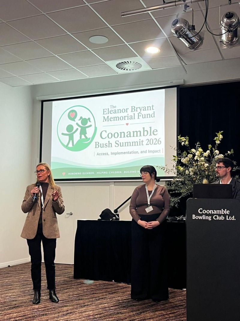 Three women are standing in front of a presentation screen at a conference. One woman is speaking into a microphone, another is looking on, and the third is standing behind a podium labeled "Coonamble Bowling Club Ltd.". The presentation screen displays the title "The Eleanor Bryant Memorial Fund Coonamble Bush Summit 2026" along with a logo of two children holding a heart and a figure. There is a large floral arrangement beside the podium.