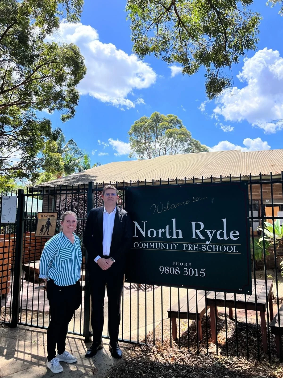 Two people standing in front of a black fence at North Ryde Community Pre-School, with a sign displaying the school’s name and phone number. The sky is blue with some clouds, and there are trees in the background.
