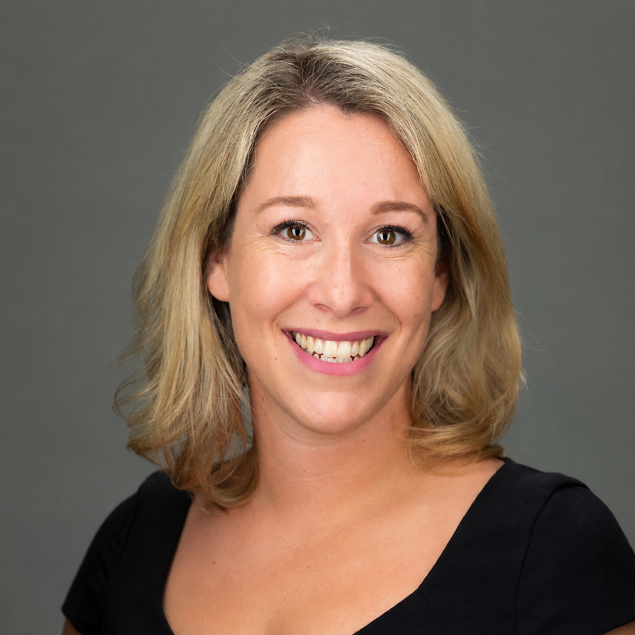 A smiling woman with shoulder-length blonde hair wearing a black top against a gray background.