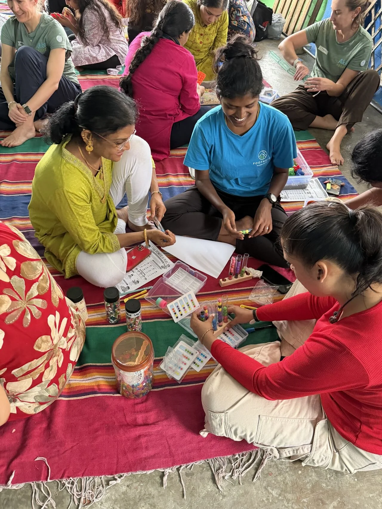 Group of women sitting on a colorful rug, engaging in arts and crafts activities with supplies like test tubes, color charts, and glitter, in an indoor setting.