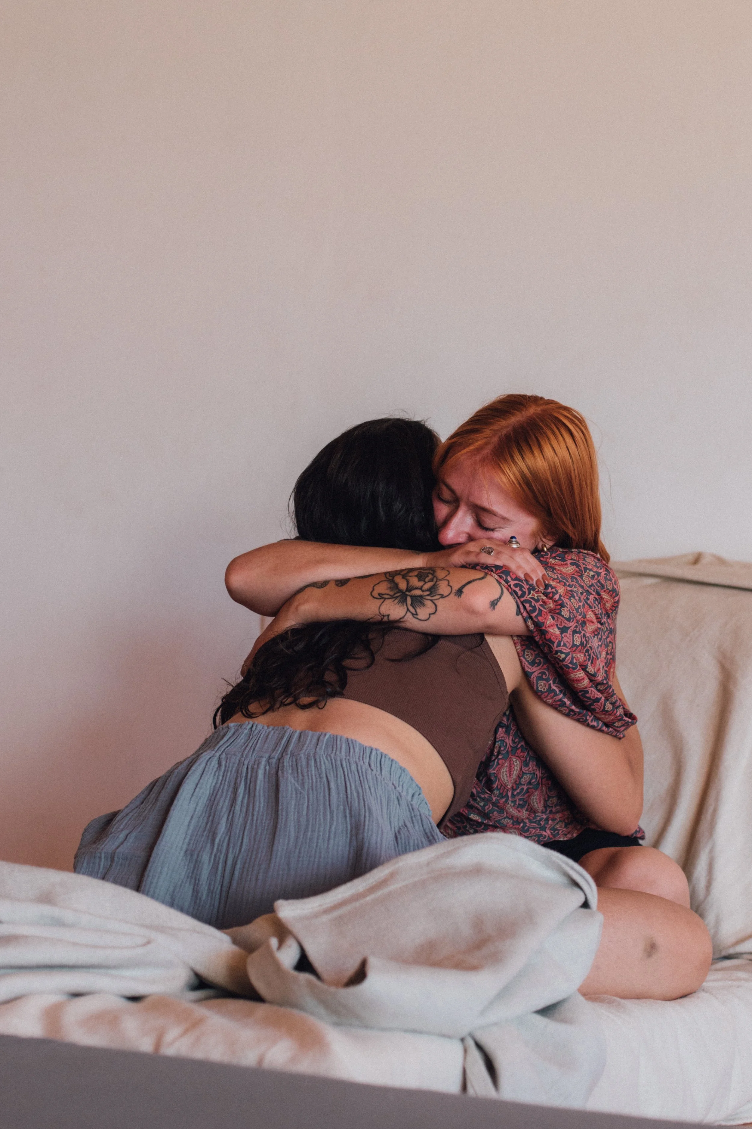 Zwei Frauen umarmen sich liebevoll in einem Schlafzimmer, eine hat rote Haare und die andere dunkle Haare, beide lächeln und zeigen Freude.