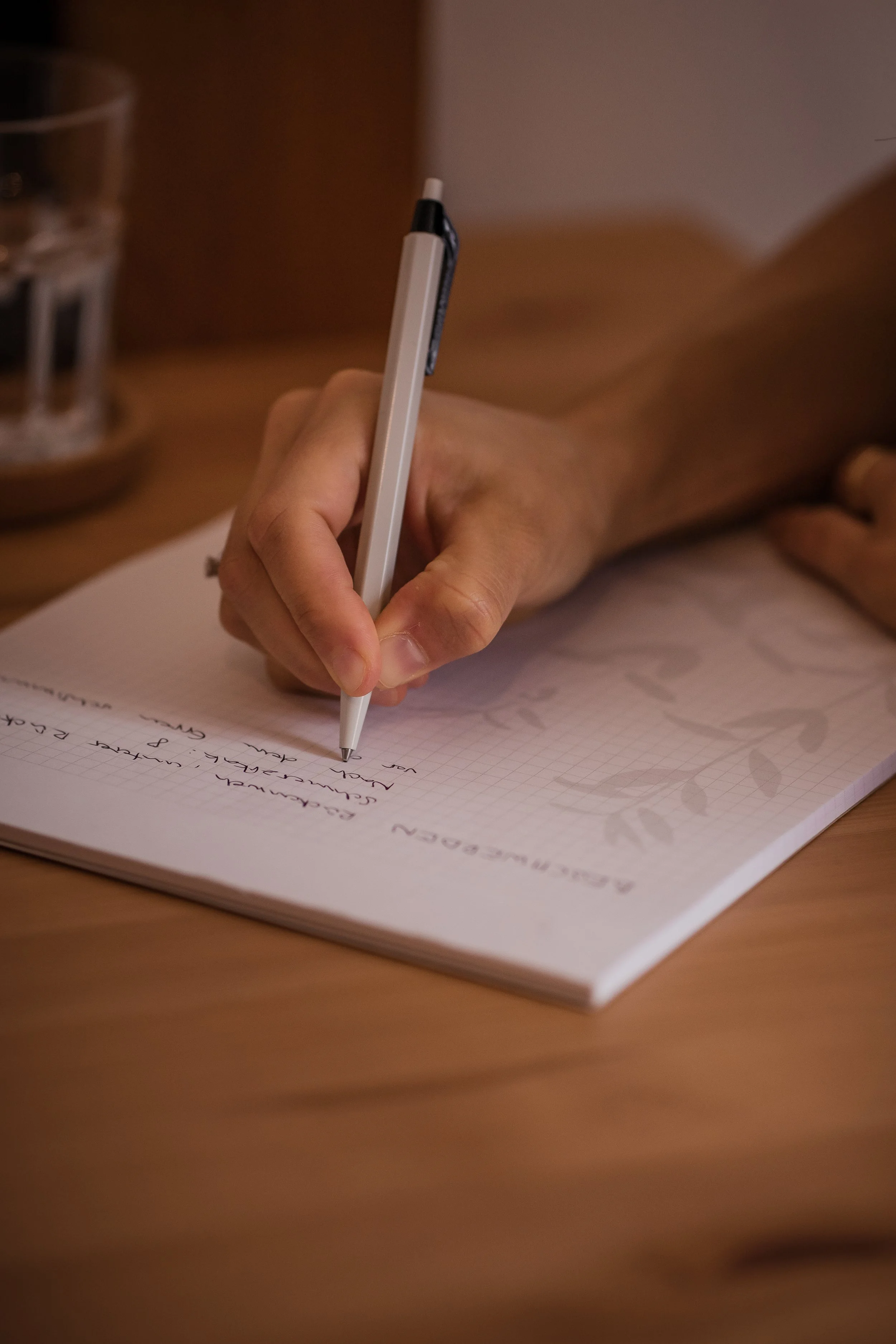 Nahaufnahme einer Hand, die in einem Notizbuch schreibt, auf einem Schreibtisch mit einem Glas Wasser im Hintergrund.