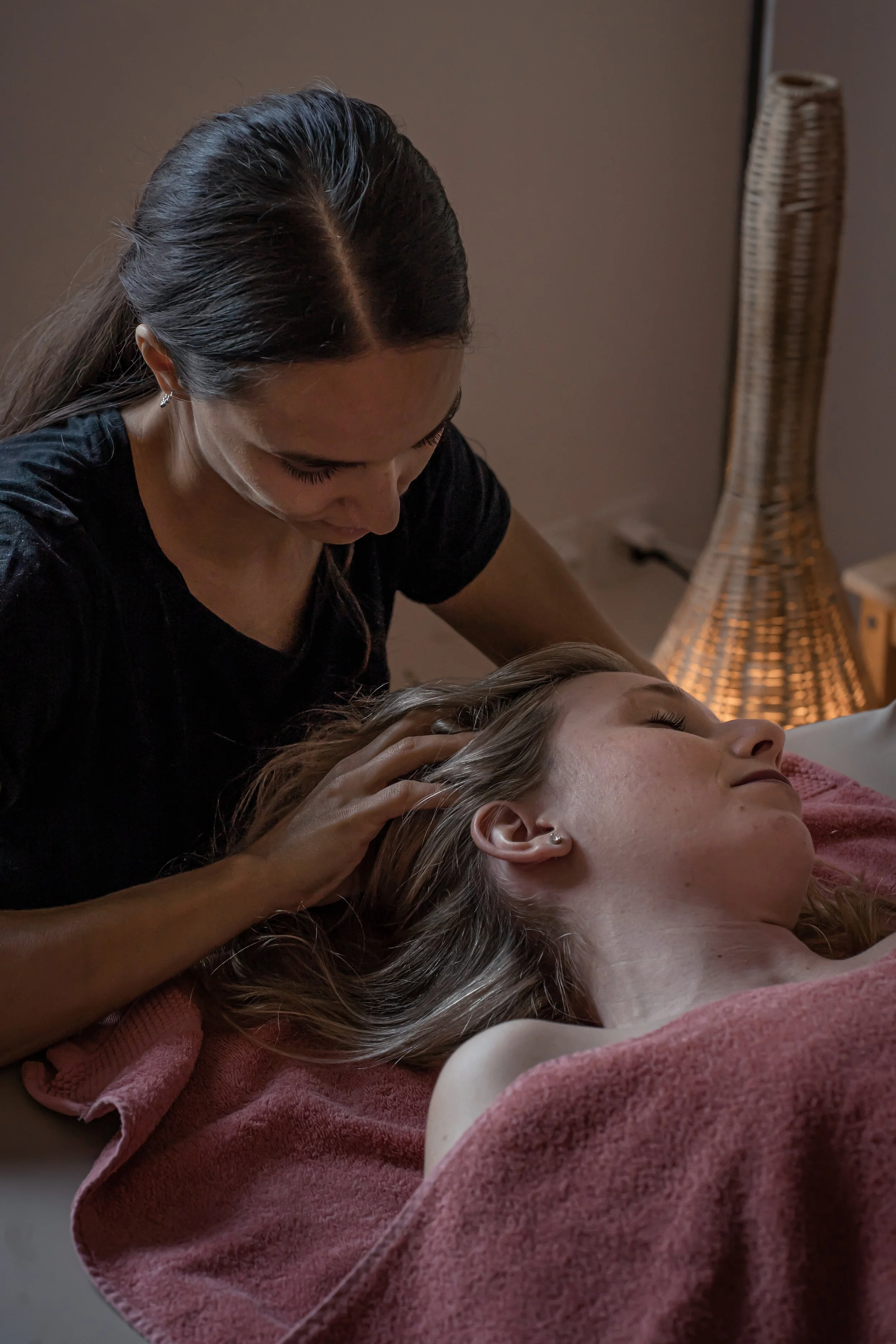 Eine Frau führt eine Massage an einem liegenden Mädchen durch, das entspannt ist. Im Hintergrund ist ein Lampenschirm.
