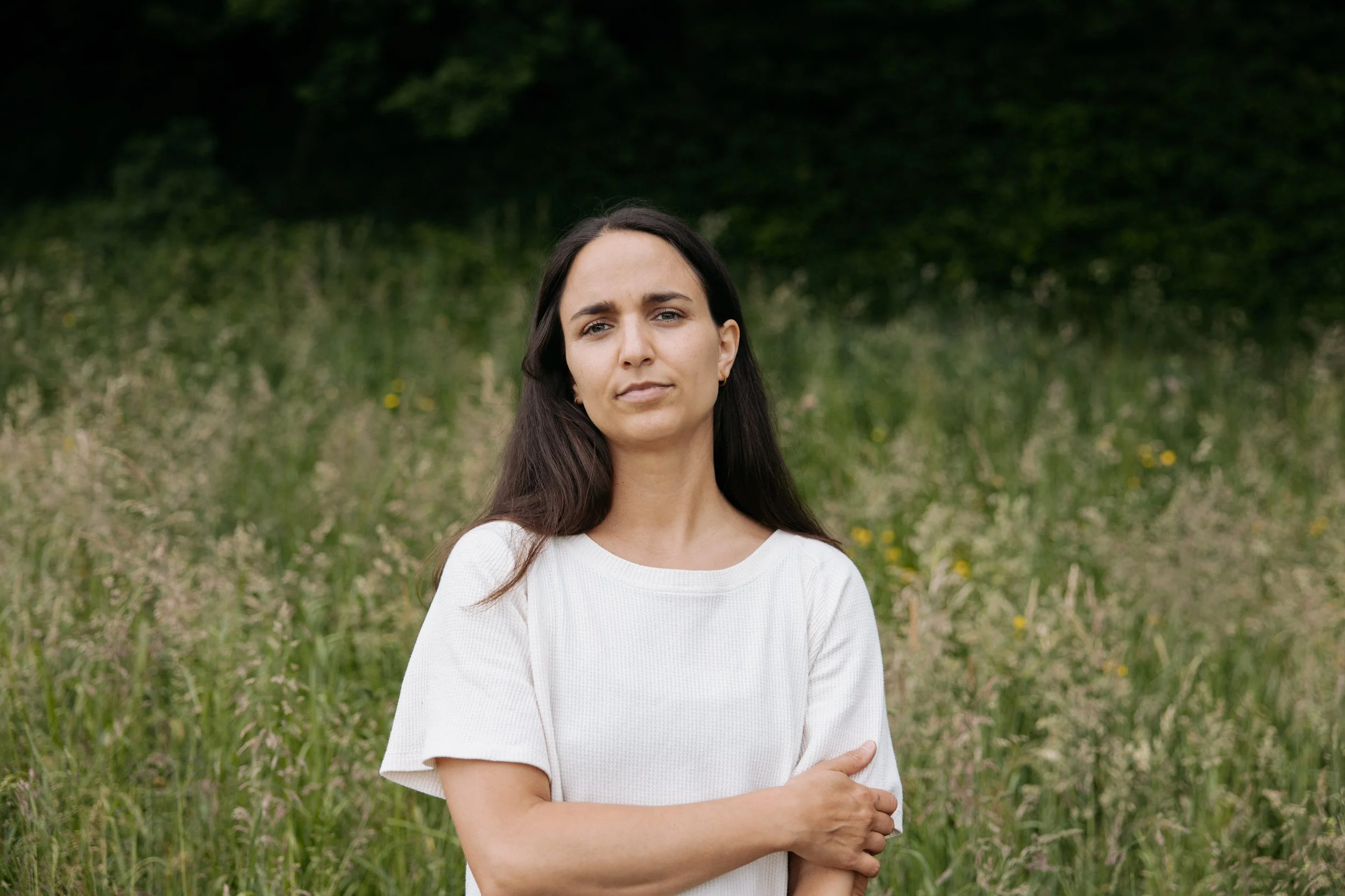 Eine Frau steht in einem grünen Feld mit Bäumen im Hintergrund, trägt ein weißes Oberteil und sieht ernst.