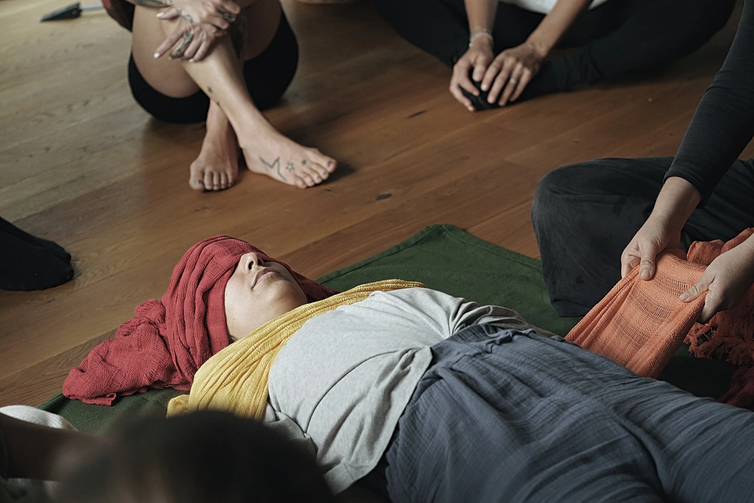 Person liegt auf dem Boden, eingewickelt in eine rote Decke, mit einem goldgelben Schal, vermutlich in einer meditativen oder entspannten Haltung, umgeben von anderen Personen im Raum. Rebozo Ritual.