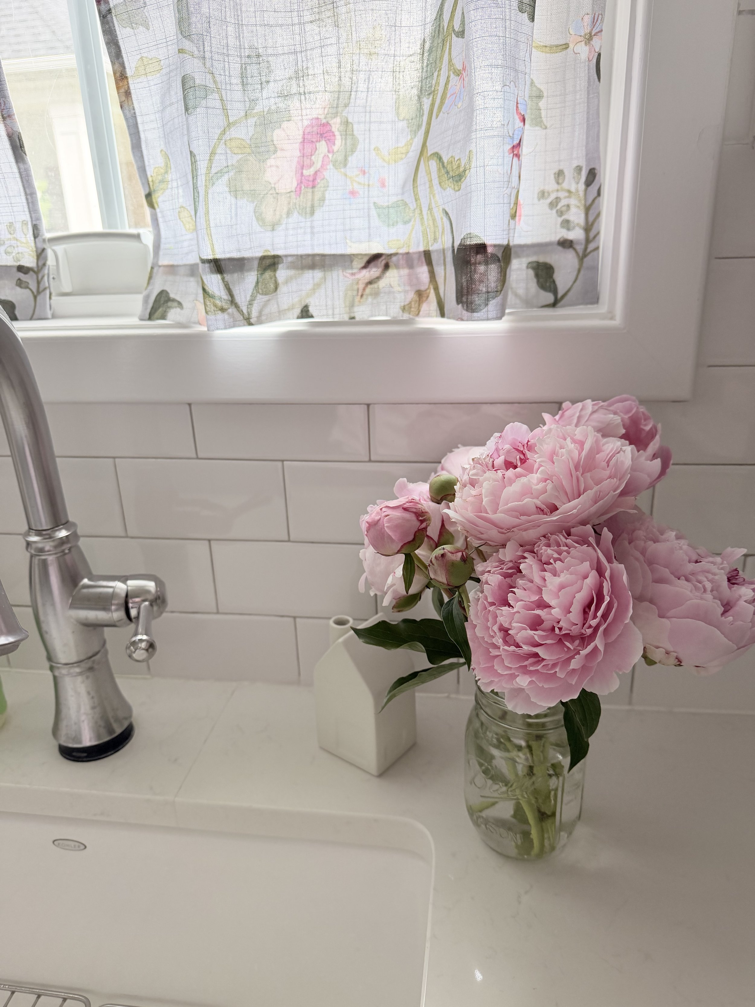 A clear glass jar holding pink peony flowers with green leaves on a white kitchen countertop near a white farmhouse sink and a chrome faucet. A box-shaped object is also on the counter, behind the flowers. A window with floral curtains is in the background.