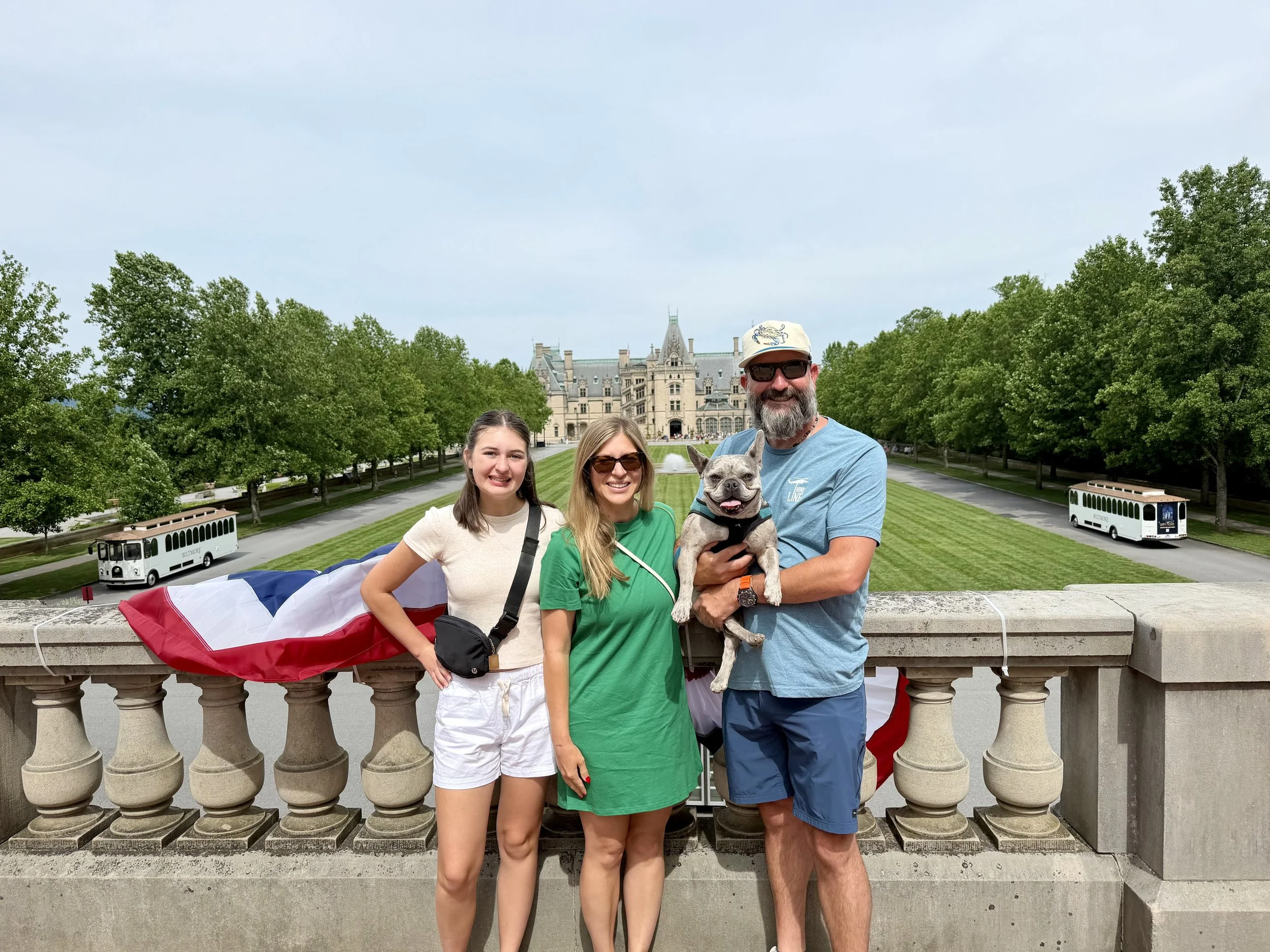 Family of four with dog standing on a bridge with castle in the background, trees, and a bus in a park or historic site.