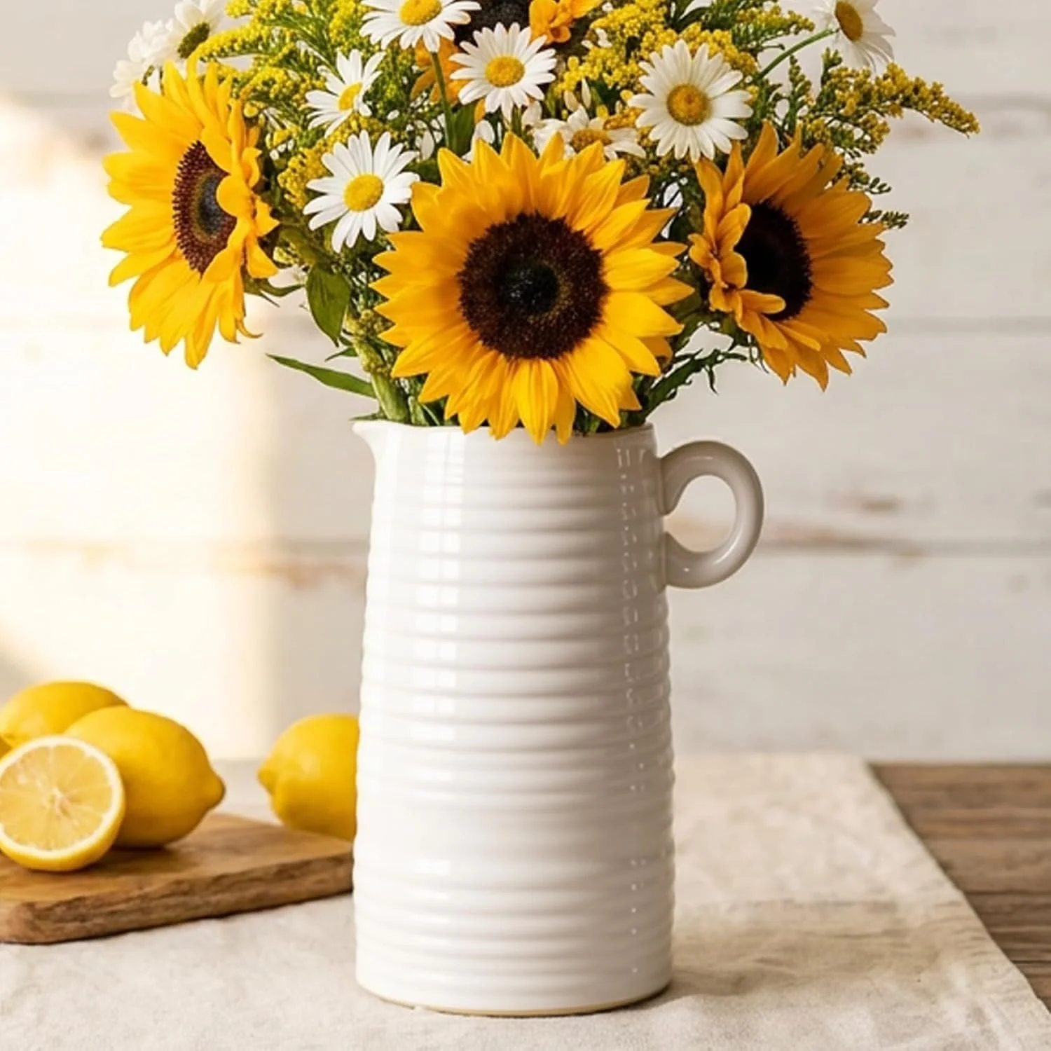 A white ceramic vase with a ribbed texture holds a bouquet of sunflowers and white daisies, set on a light-colored table with lemons nearby.