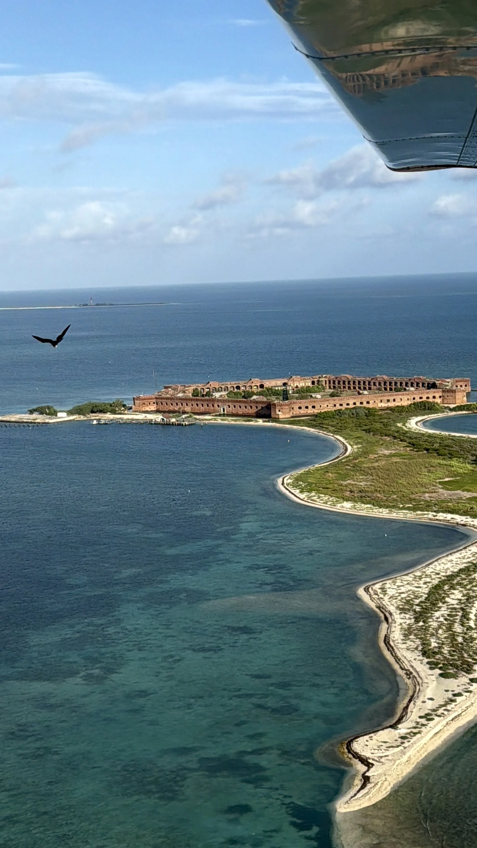Aerial view of an island with a large historic fortress or castle, surrounded by water, next to a narrow strip of beach or sand, with a bird flying in the sky and a trail of land in the distance.