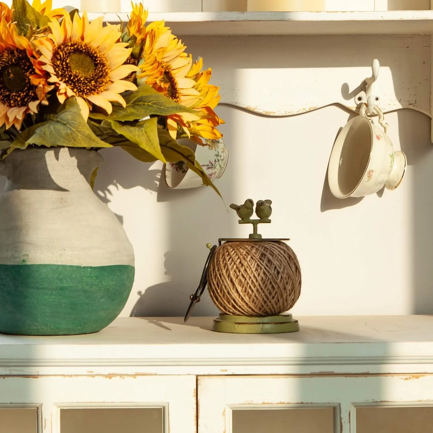 A large ceramic vase with a two-tone paint gradient from beige to green, holding a bouquet of sunflowers and green leaves. On the shelf behind, a small floral teacup with a saucer hangs from a hook. On the surface, there's a vintage-style wooden spool of brown twine with a small bird-shaped ornament on top and a pair of scissors.