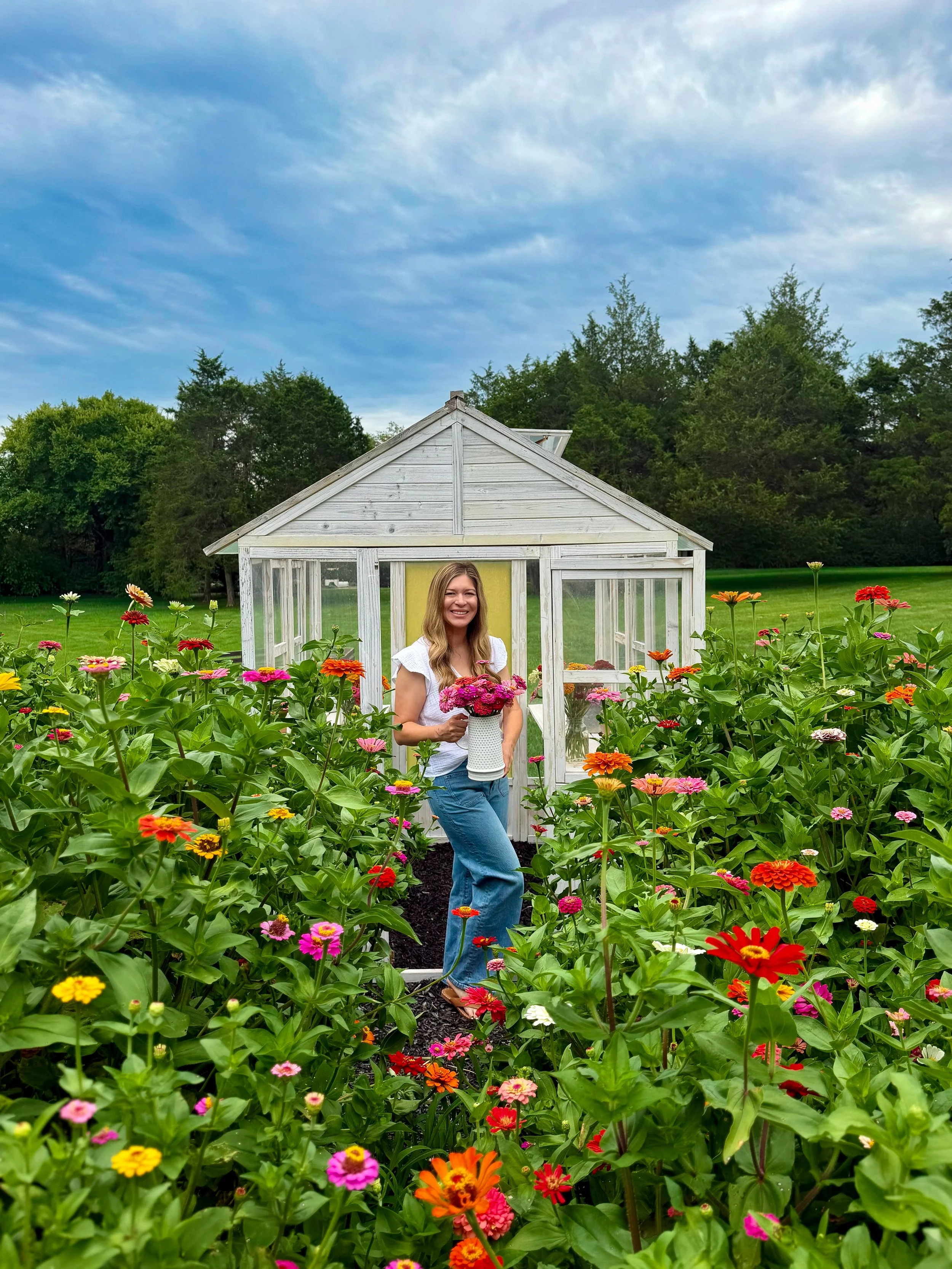 A woman standing among colorful zinnia flowers in front of a small white greenhouse with a yellow door, surrounded by green trees and grass under a partly cloudy sky.