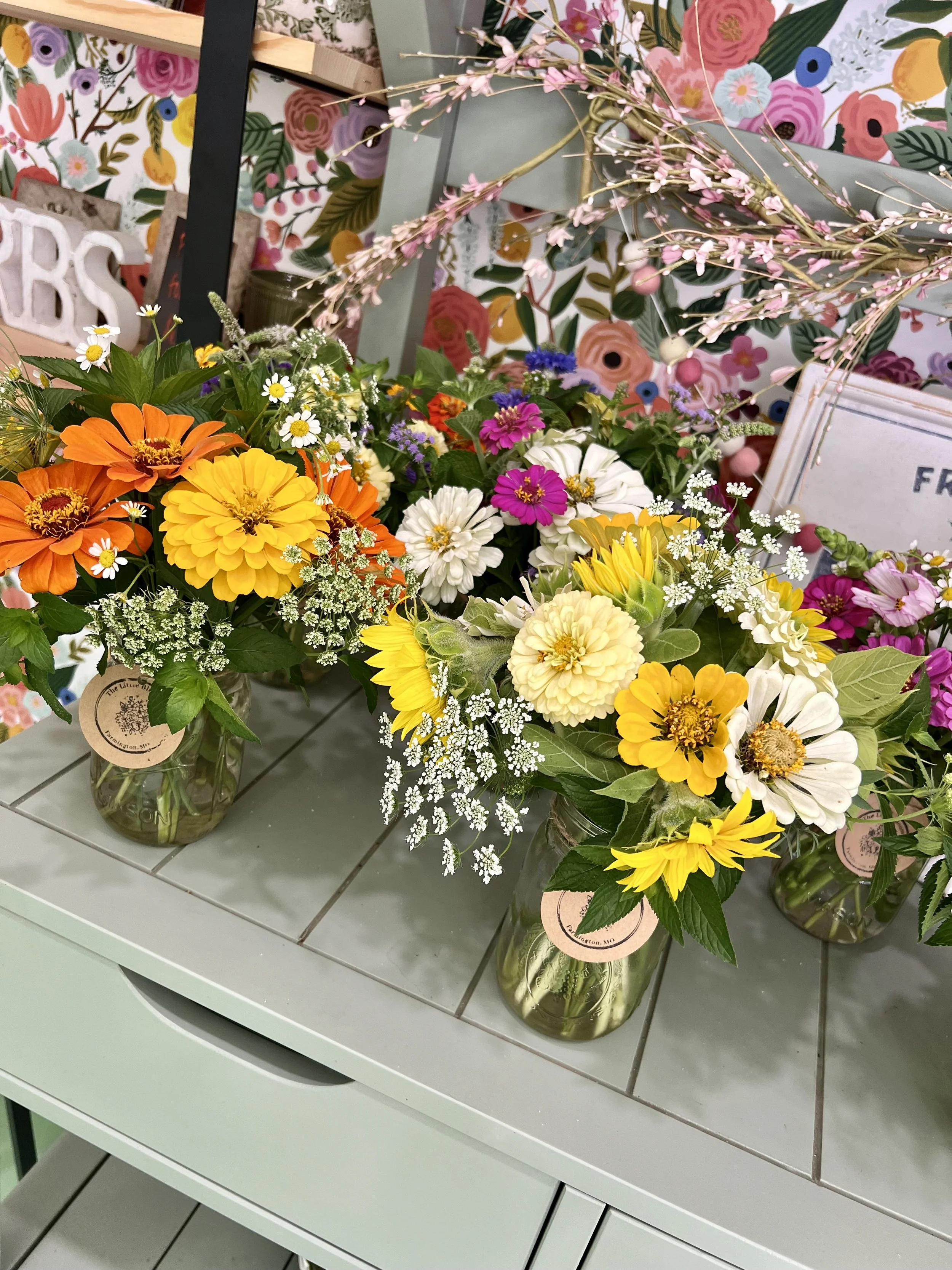 Vases filled with colorful flowers such as sunflowers, daisies, and other mixed floral arrangements on a white surface with a floral wallpaper background.