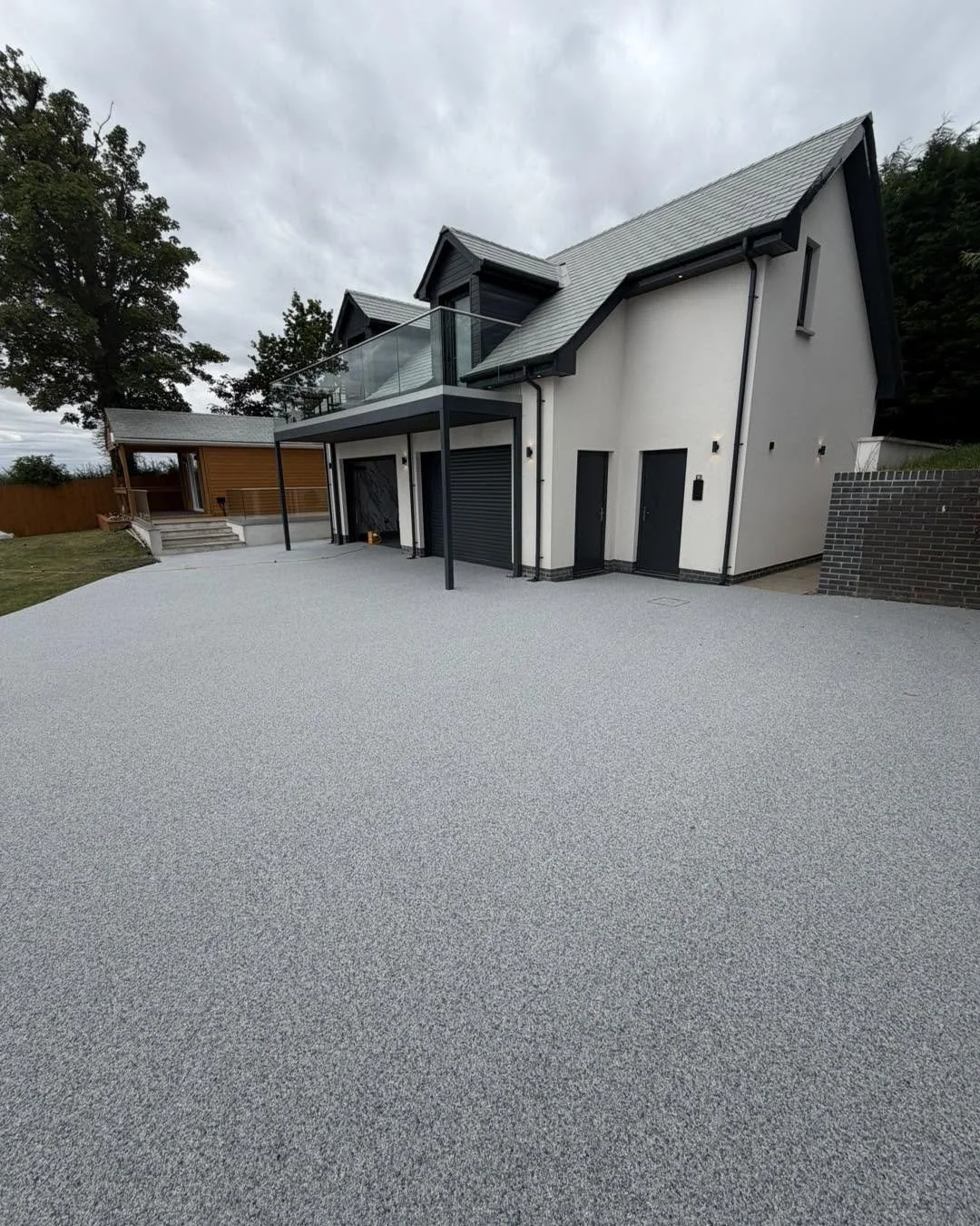 Modern house with a textured concrete patio, black metal accents, and a glass balcony on the second floor under a cloudy sky.