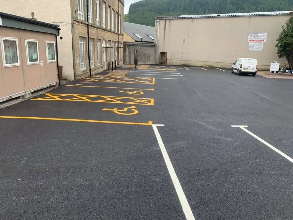 Empty parking lot outside a building with designated parking spaces marked with yellow and white lines. A white van is parked toward the back, and there are signs posted on the building and to the right side of the lot.