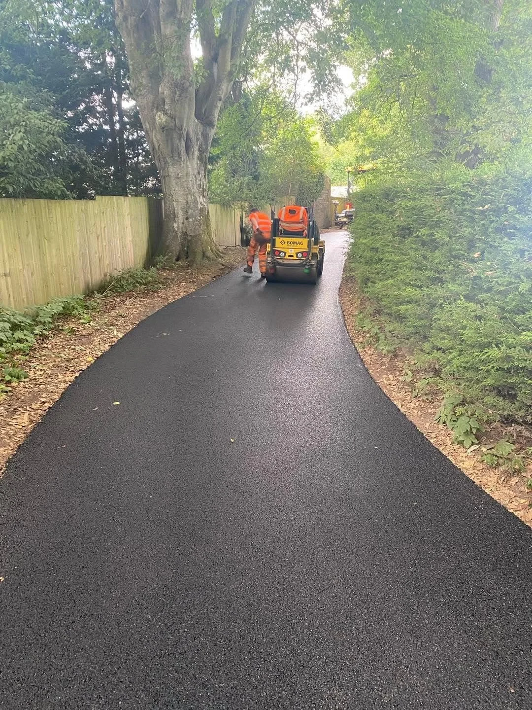 Construction workers in orange safety vests and helmets working on a freshly paved asphalt pathway in a wooded area, with a Bamac compact roller machine nearby.