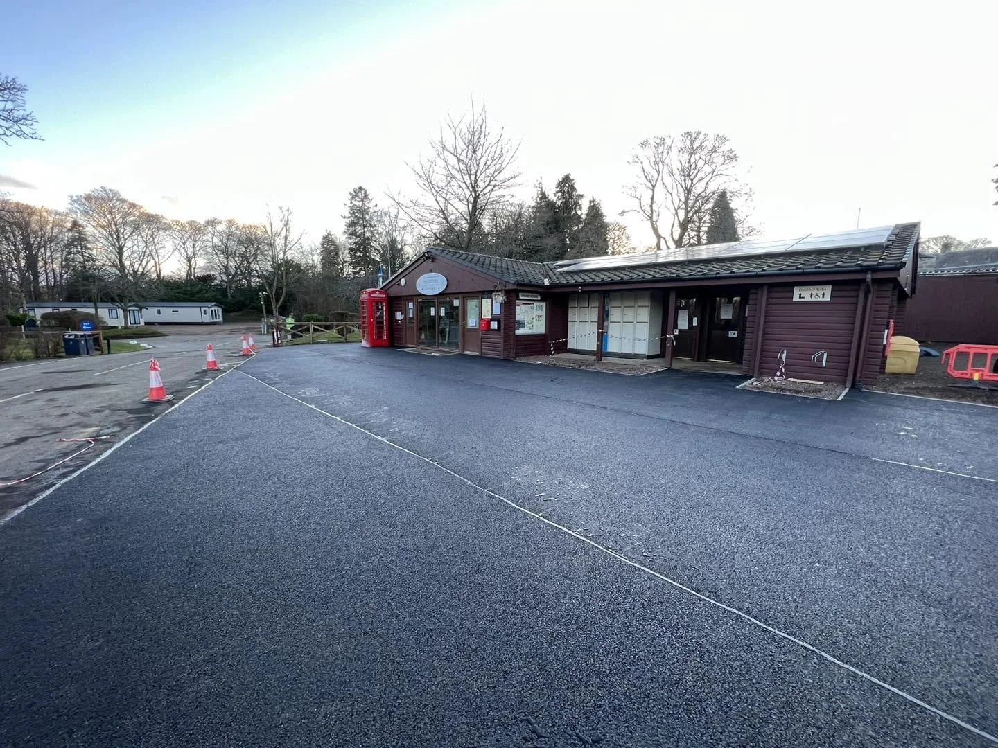 A small wooden building with a dark brown exterior and a tiled roof, situated in a quiet, rural area. There is a red telephone booth next to the building, with orange traffic cones and a cord indicating construction or maintenance work nearby. Bare trees are in the background under a clear sky.