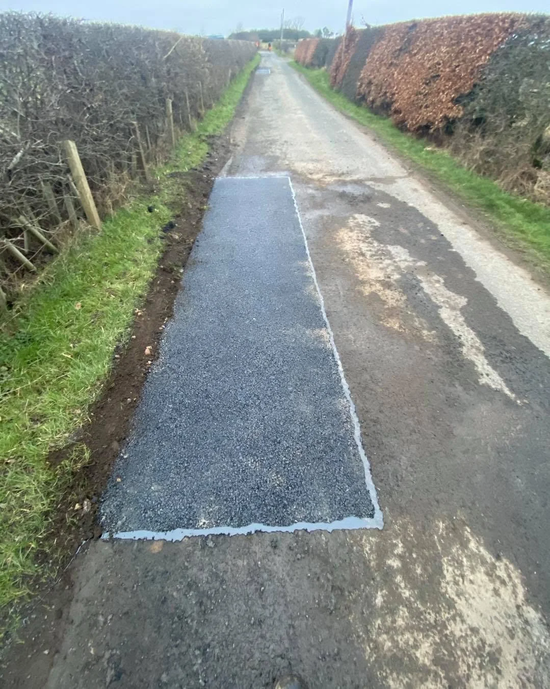 A section of freshly paved asphalt on a rural road, with a white outline marking the edges of the new pavement. The road is flanked by bushes and grass on either side.
