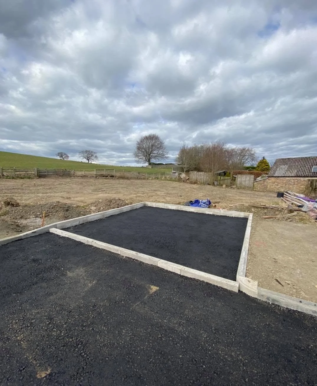 A construction site with a newly built black asphalt driveway and a gravel area, surrounded by a rural landscape with trees, fences, and houses under a cloudy sky.