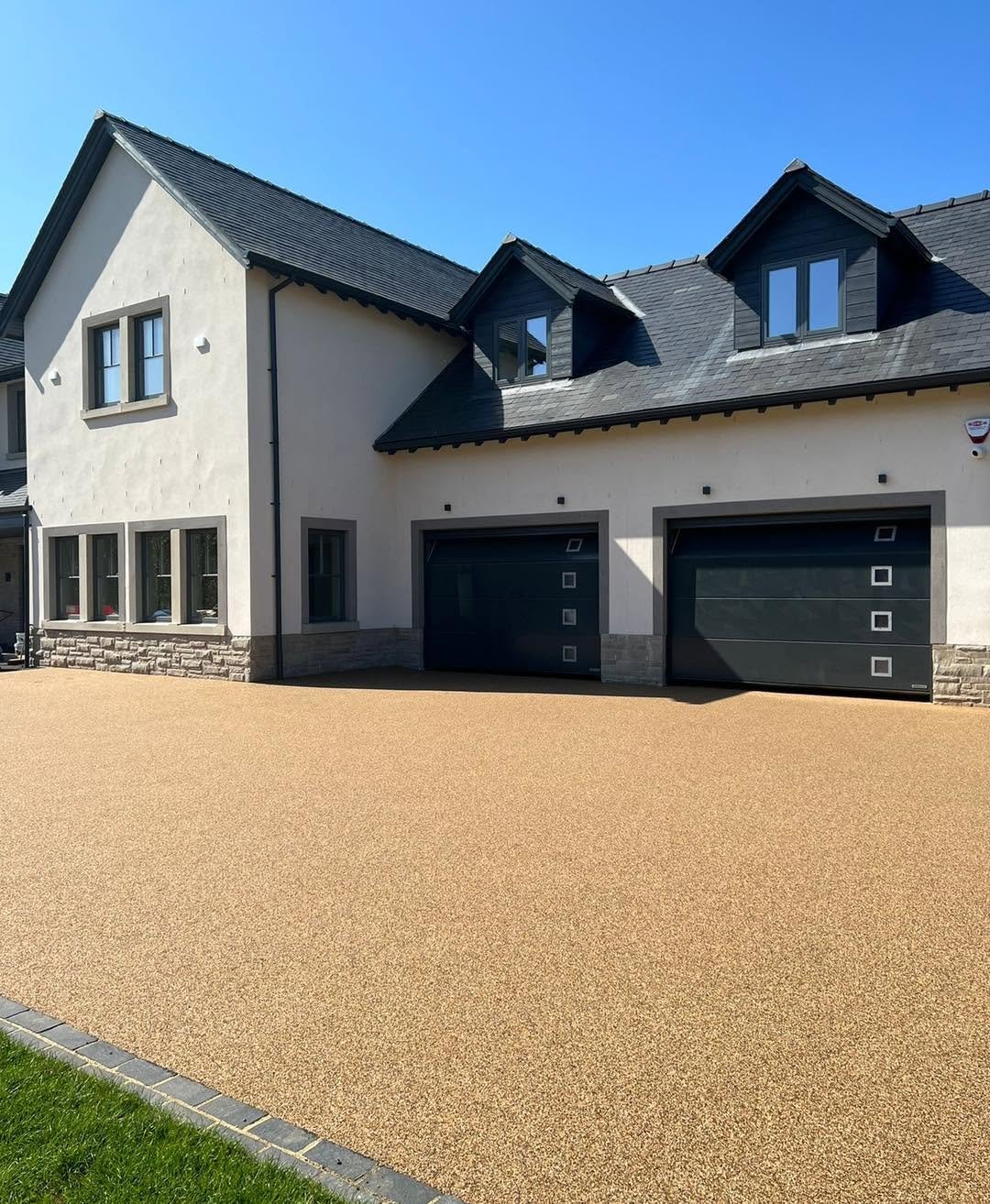 Modern house with a pitched roof, garage doors, and a spacious front yard with a textured beige driveway and a small strip of grass.