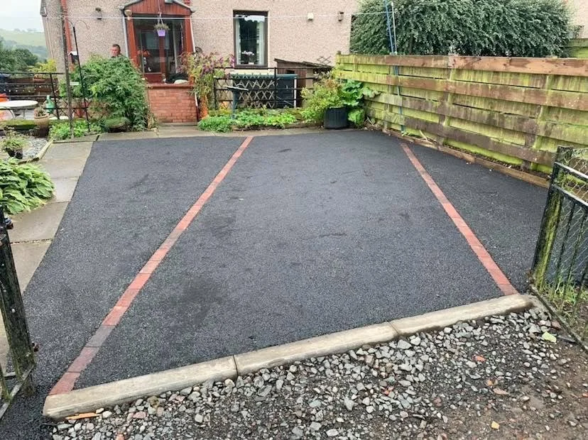 Empty parking space with asphalt, separated by red bricks, surrounded by a small gate and garden at the back of a house.