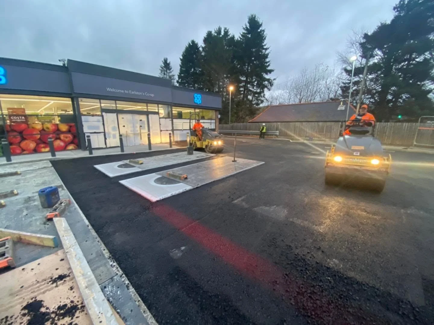 Workers are paving the parking lot outside a grocery store at dusk, with one person operating a roller and another on a ride-on machine.