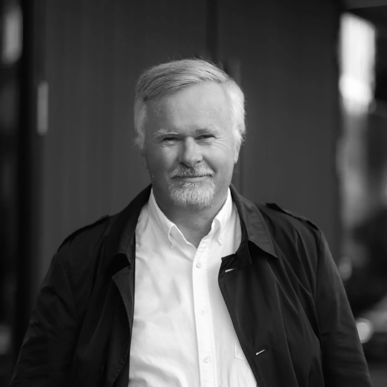 Black-and-white photo of a smiling middle-aged man with gray hair and beard, wearing a white shirt and dark jacket, standing indoors.