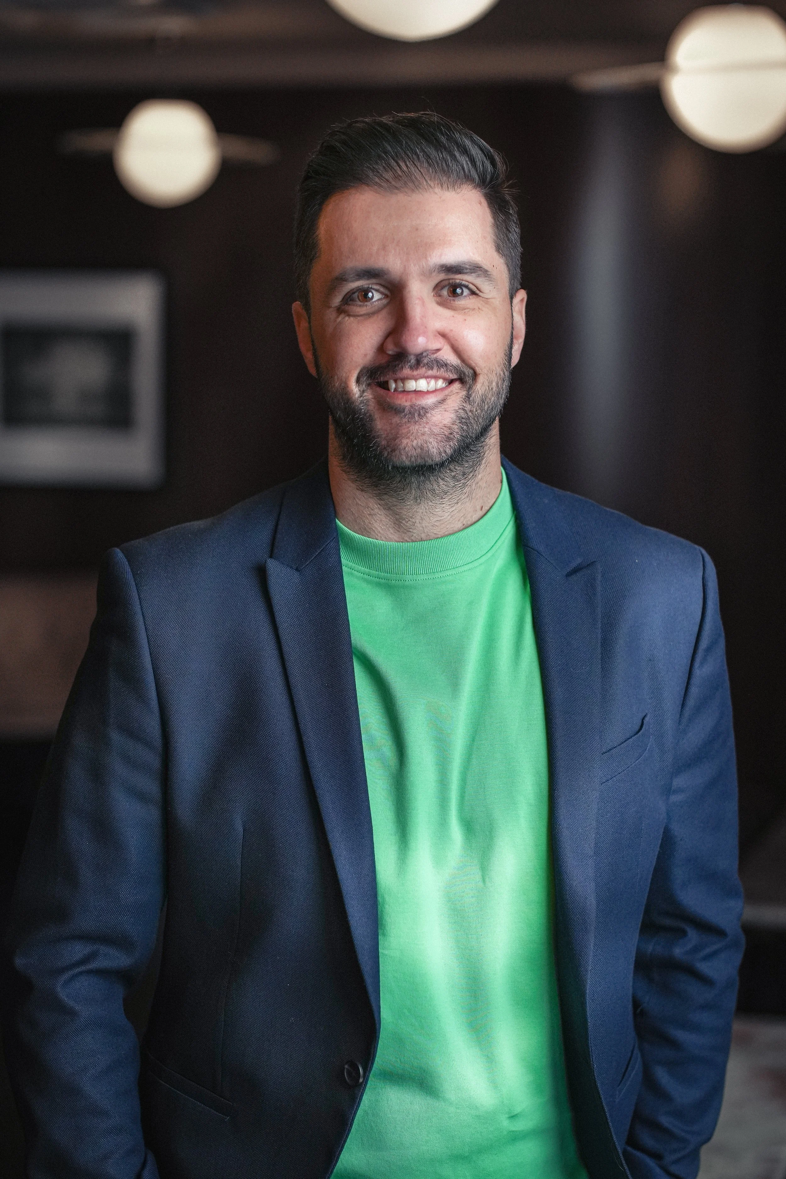 Adrian Diaz, a man with dark hair, a beard, and brown eyes smiling, wearing a dark blue blazer and a bright green t-shirt, standing indoors with a dark background and round ceiling lights.