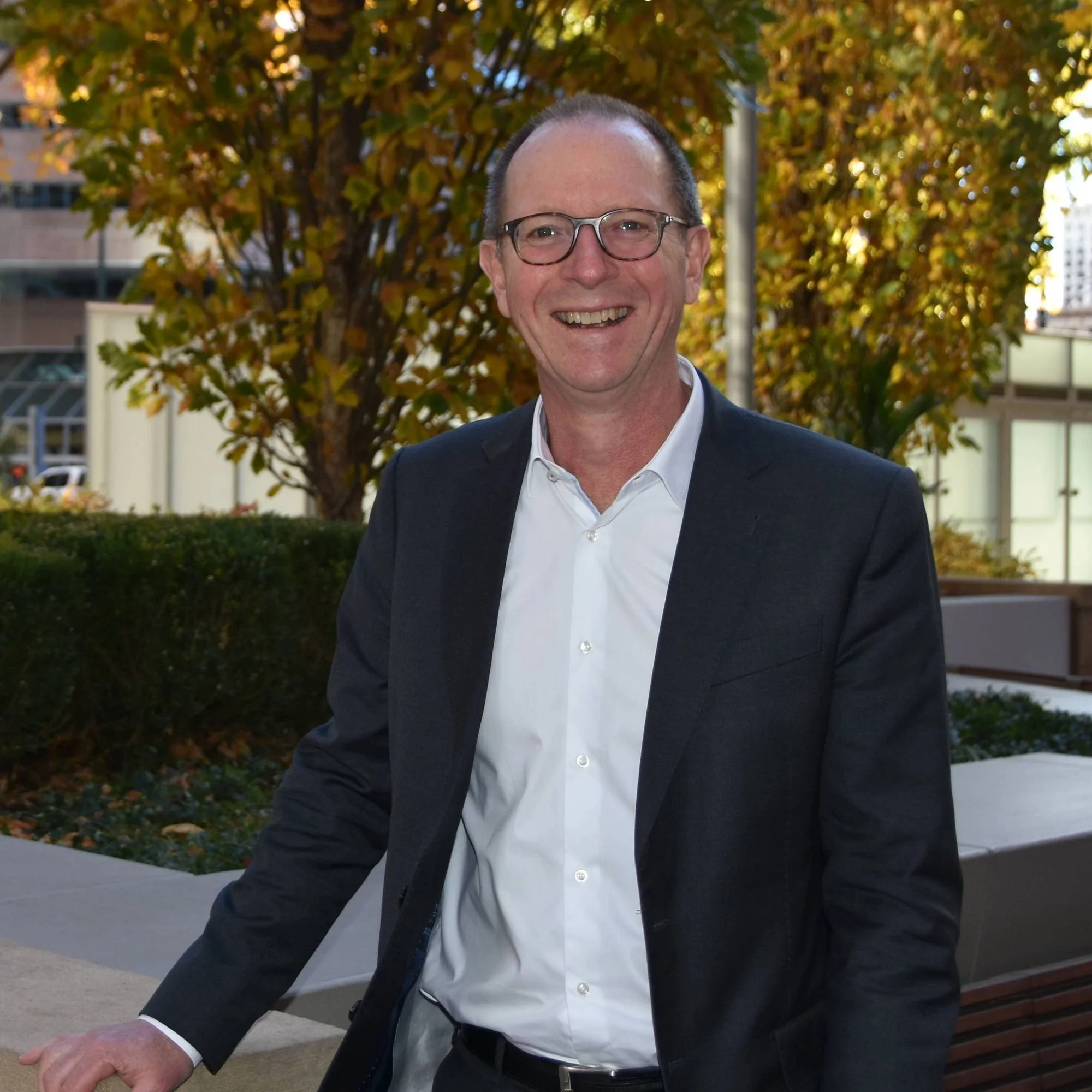 A smiling middle-aged man wearing glasses, a white shirt, and a dark blazer standing outdoors in front of autumn-colored trees, with buildings and a sidewalk in the background.
