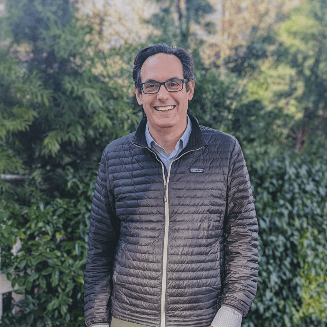 A man with glasses smiling outdoors in front of green foliage, wearing a black puffy jacket over a light blue collared shirt.