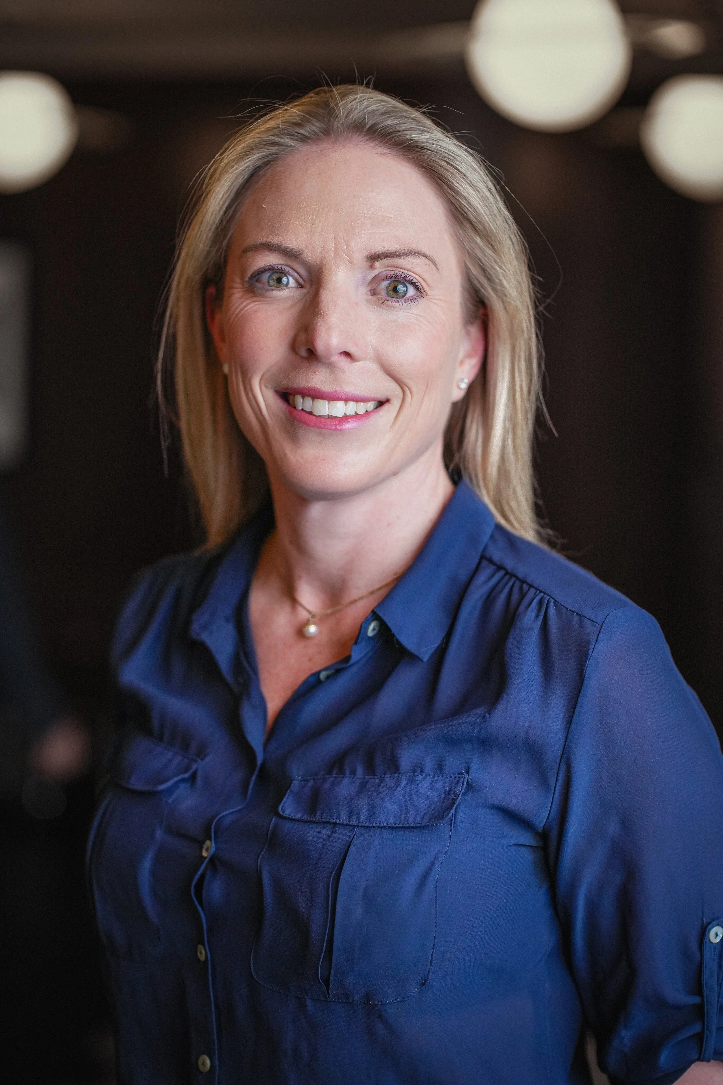A woman with blonde hair and blue eyes smiling. She is wearing a blue shirt and a pearl necklace. The background is blurred with white circular lights.