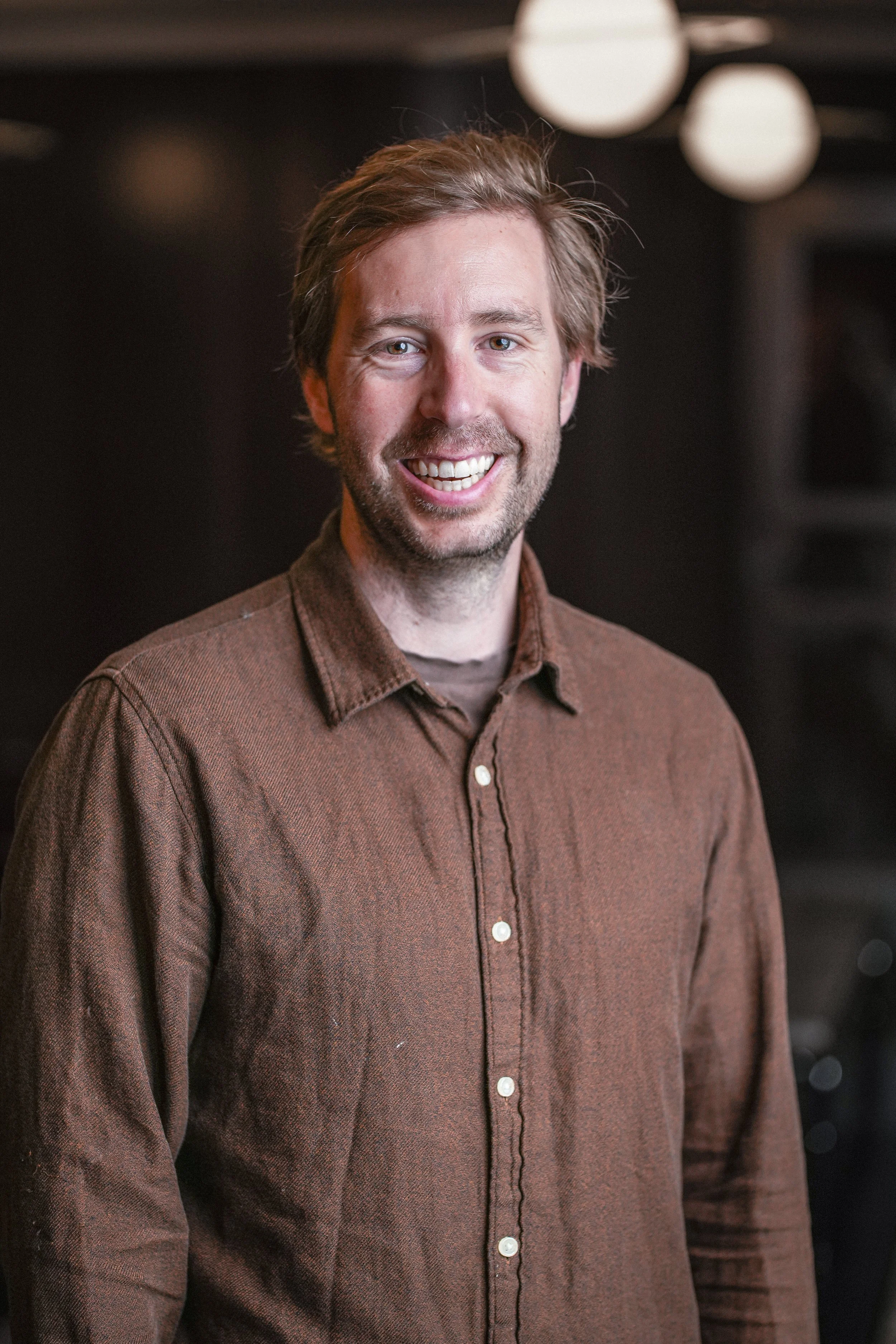 Engebreth Faerden, a smiling man with light brown hair and a beard, wearing a brown button-up shirt, standing indoors with lights in the background.