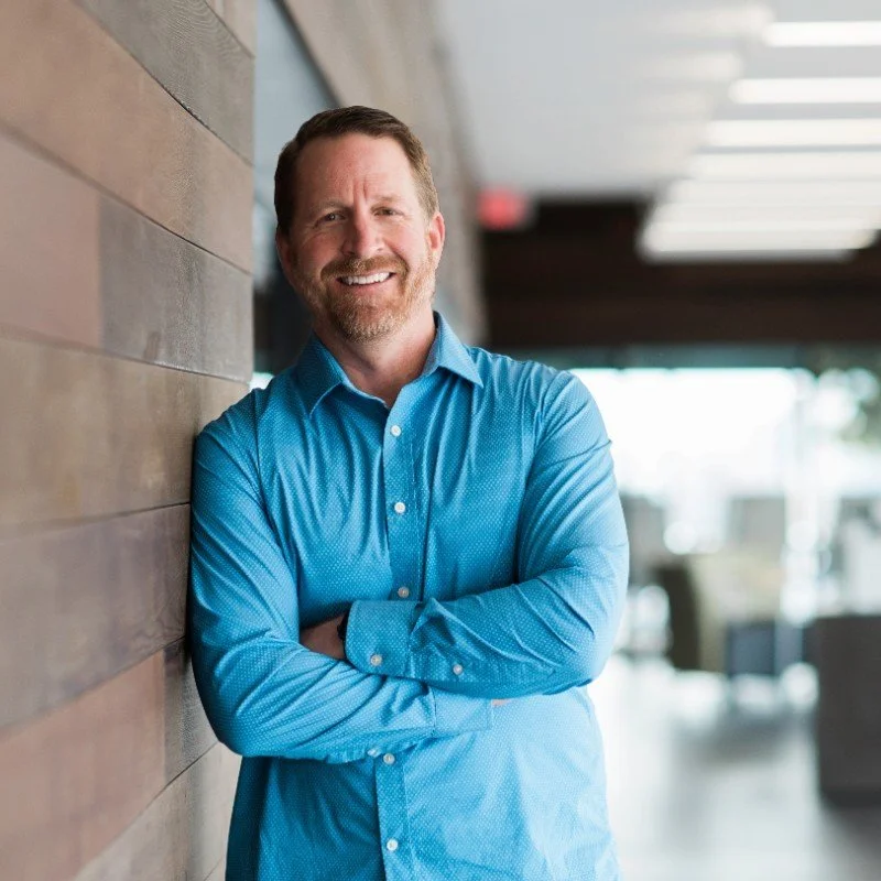 A man with a beard in a blue dress shirt leaning against a wooden wall, smiling indoors with a modern background.