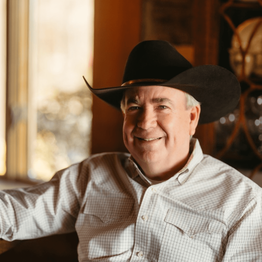 A smiling man wearing a cowboy hat and a checkered shirt, sitting indoors near a window.