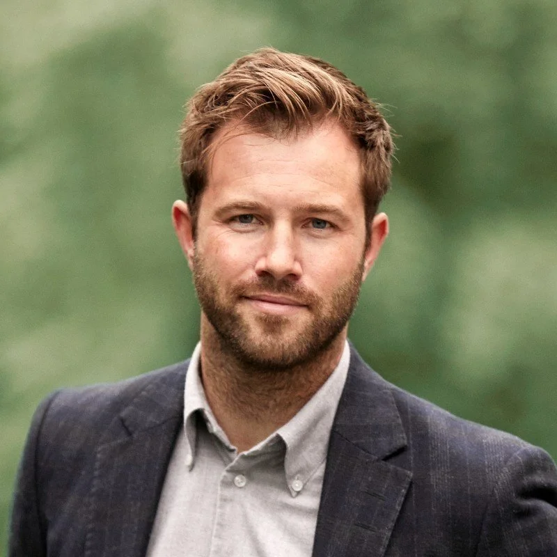 A man with light brown hair and a beard, dressed in a gray blazer over a light gray shirt, standing outdoors with a blurred green background.