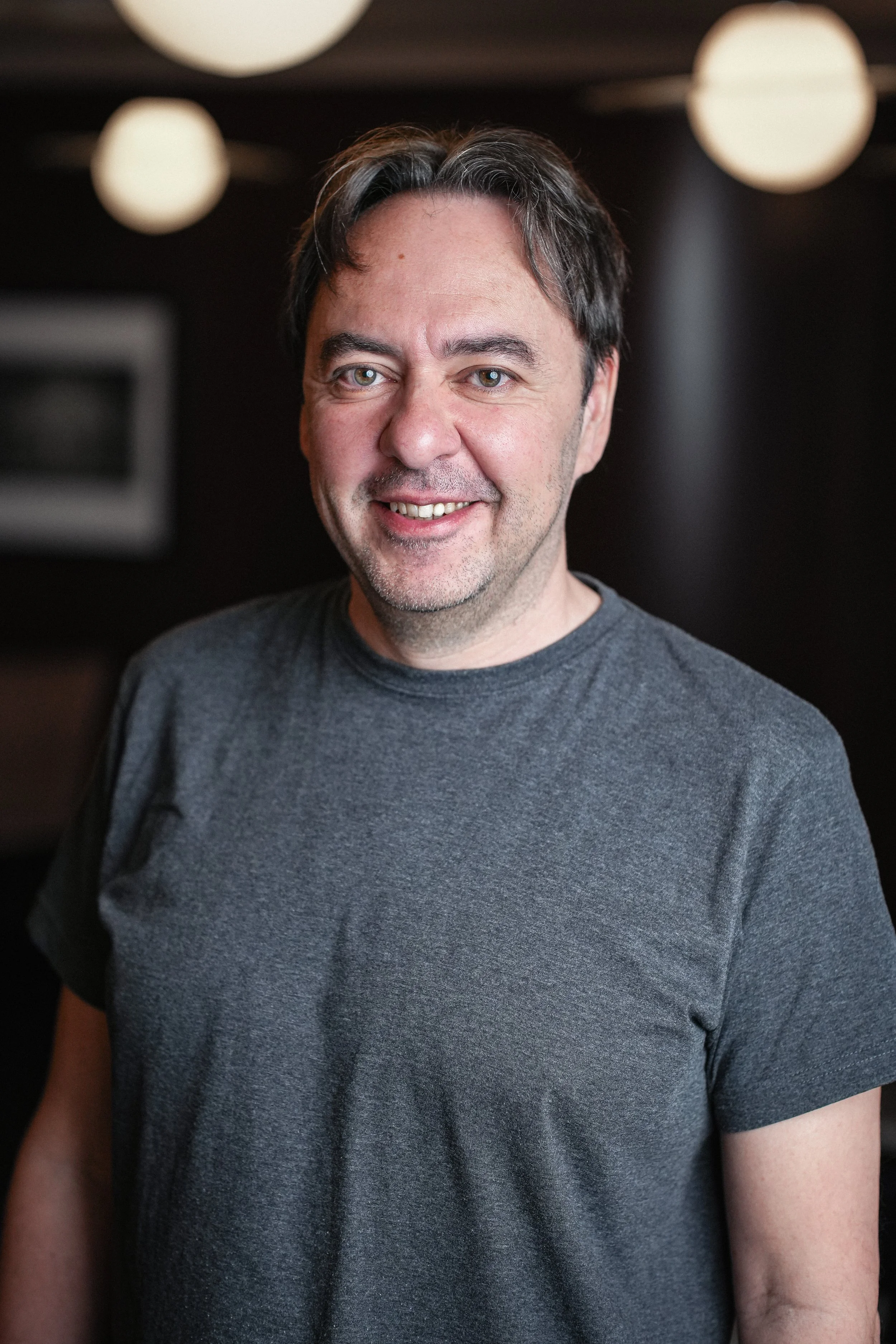 David Moriano, a man with short dark hair, blue eyes, smiling, wearing a gray t-shirt, standing in a room with soft lighting and round ceiling lights.