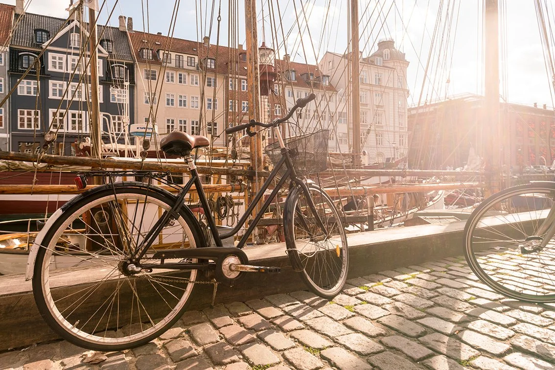 A black bicycle parked on a cobblestone pathway next to a harbor with sailboats and colorful buildings in the background, sunlight shining brightly.