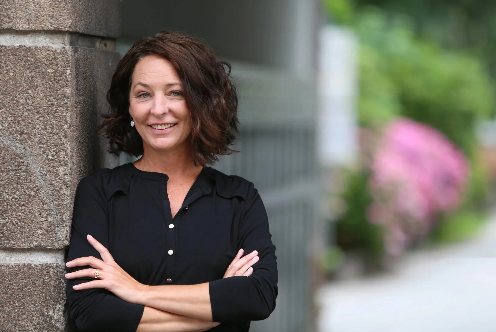 A middle-aged woman with curly brown hair stands outdoors, smiling, with her arms crossed, leaning against a brick wall.