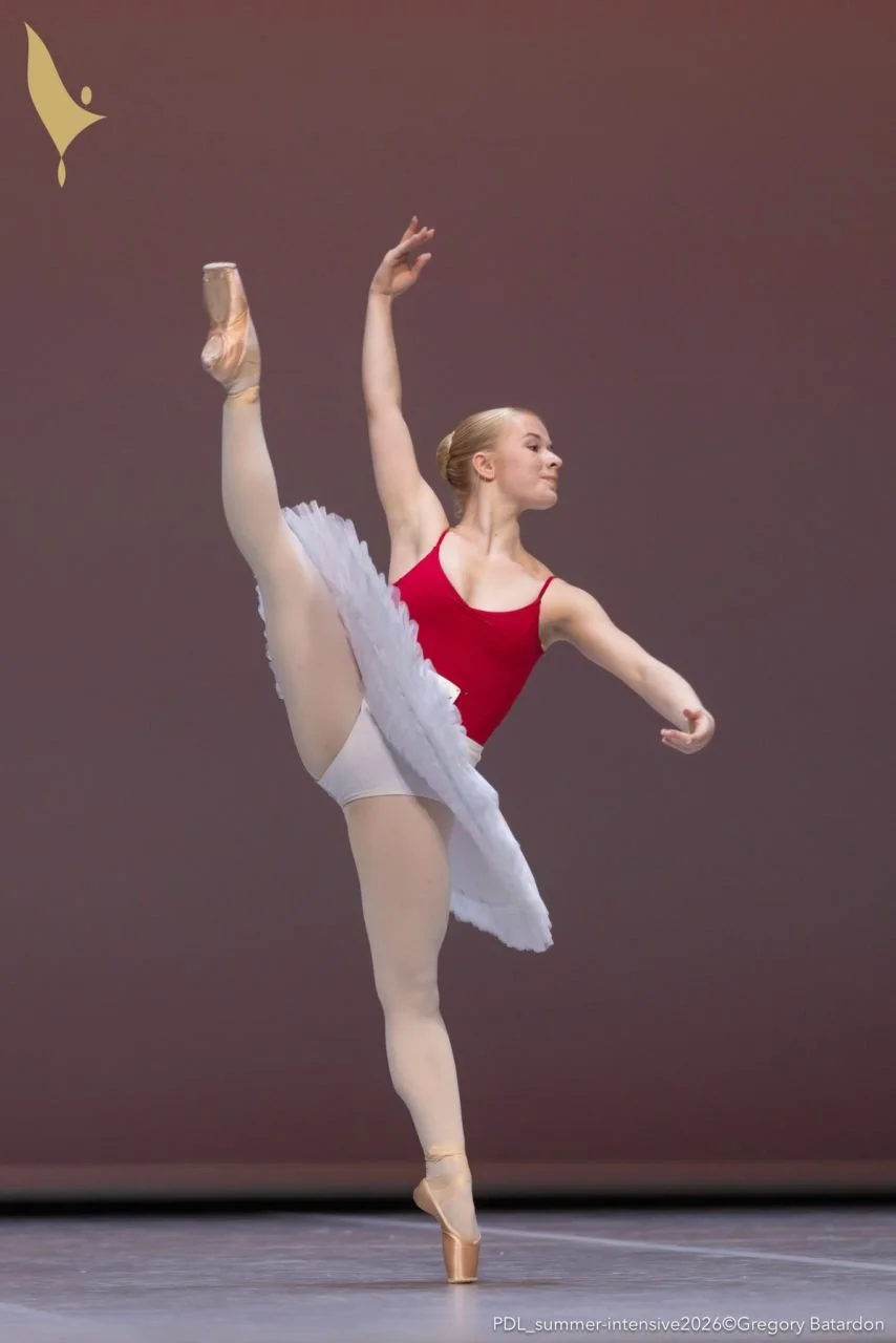 Ballet dancer en pointe performing a high leg extension in a red leotard and tutu.