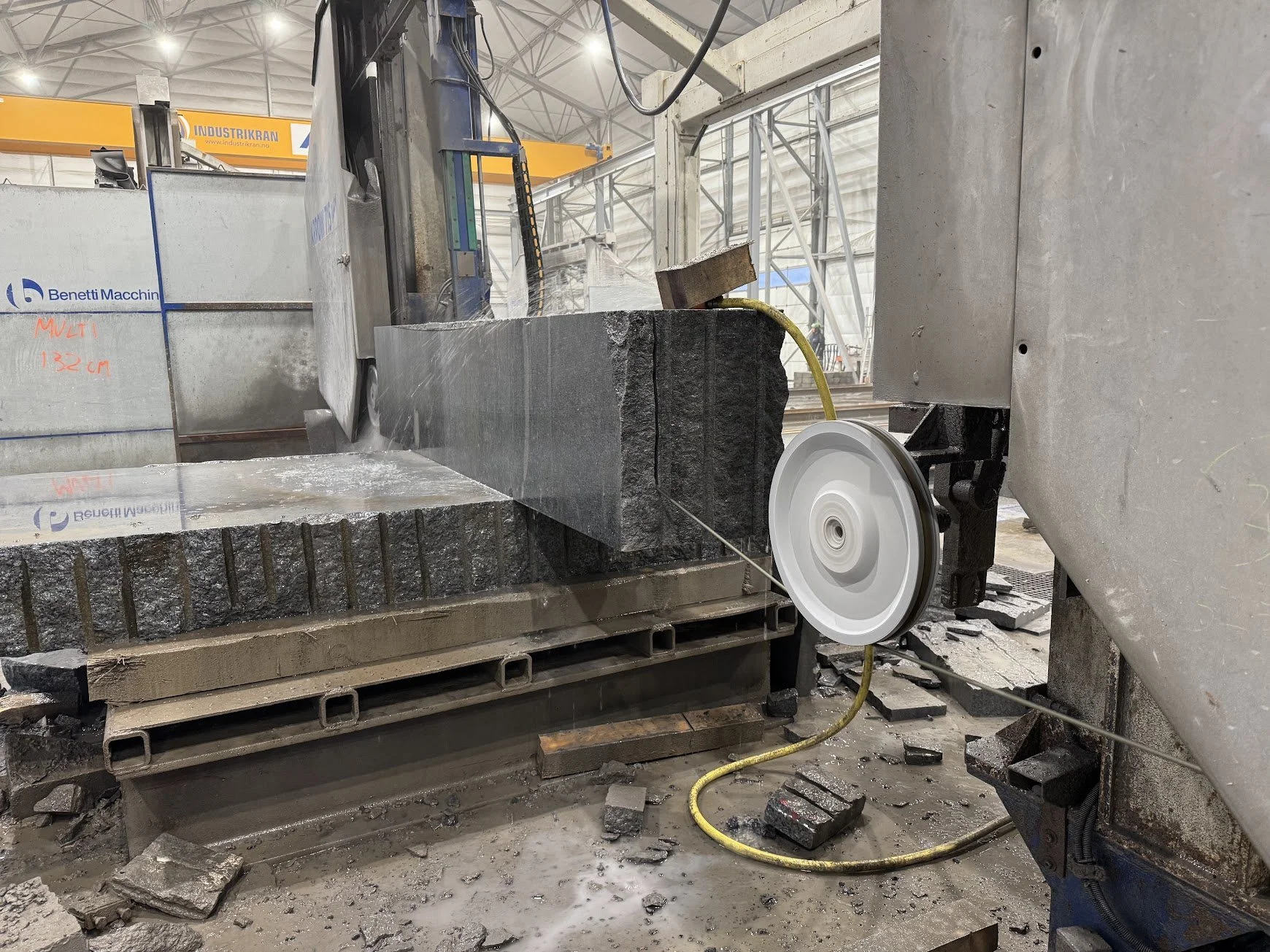 En industriell maskin som skjærer eller skår opp stein i en fabrikk eller verksted, med steinbiter på gulvet og kontrollpaneler på maskinen.