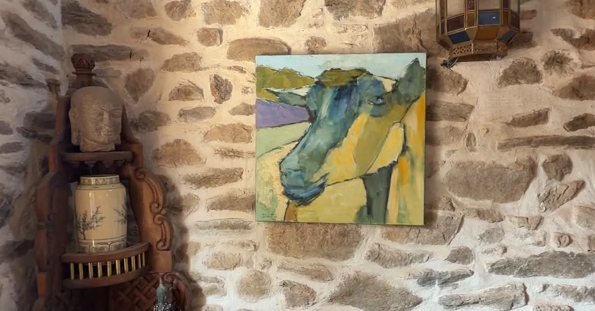 A colorful painting of a cow hanging on a stone wall. To the left, there's a wooden shelf with a decorative sculpture and a vase, and to the right, part of a patterned lantern is visible.