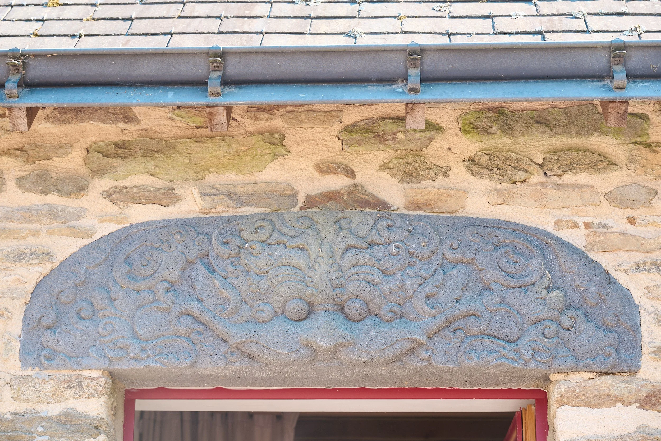 Close-up of a building's exterior featuring a stone wall with a carved decorative panel, a metal gutter, and a partially opened window with a red frame and orange curtain.