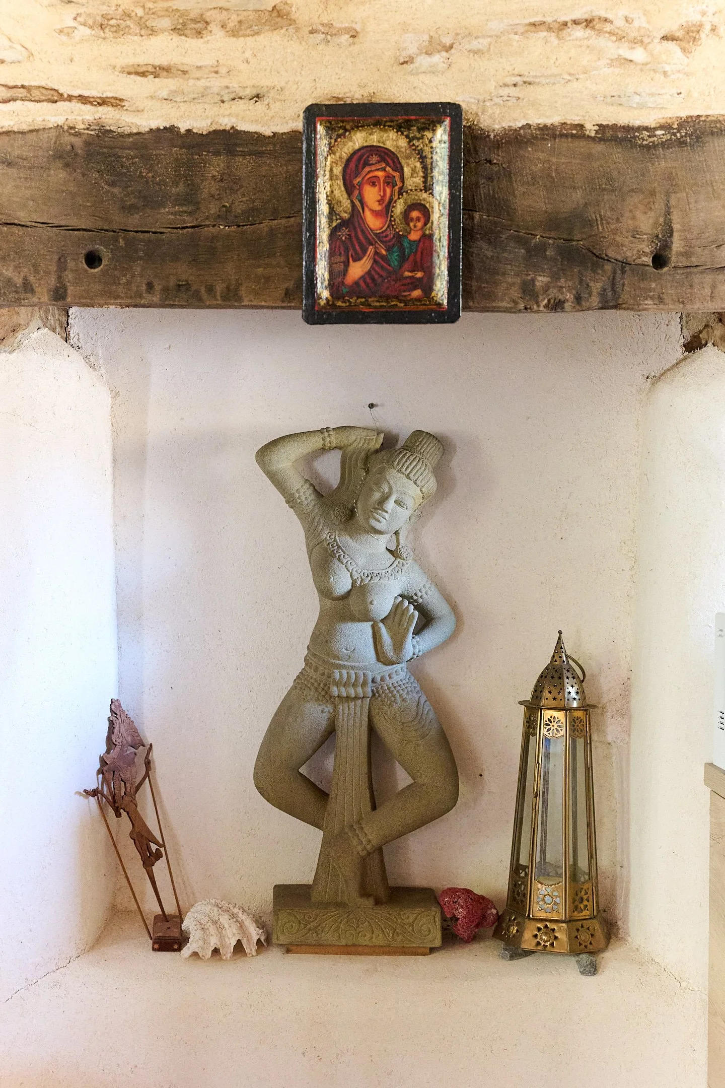 Decorative display featuring a carved stone sculpture of a woman holding a jar, a small religious icon of the Madonna and Child above, and a brass lantern on the right. Other smaller items are on the shelf below.
