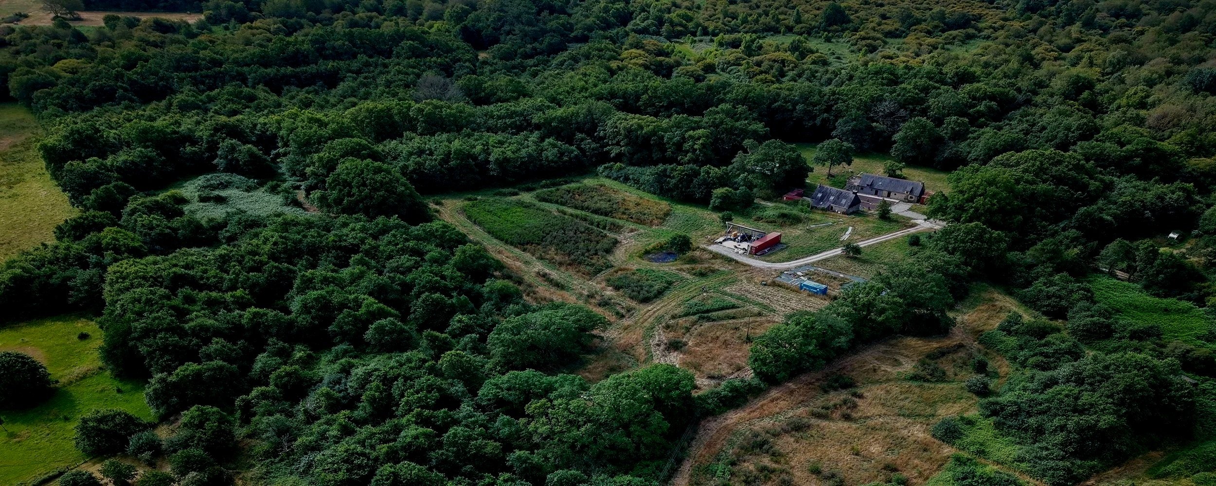 Aerial view of a rural farm with cultivated fields, trees, a house, barns, and farm equipment surrounded by lush greenery.